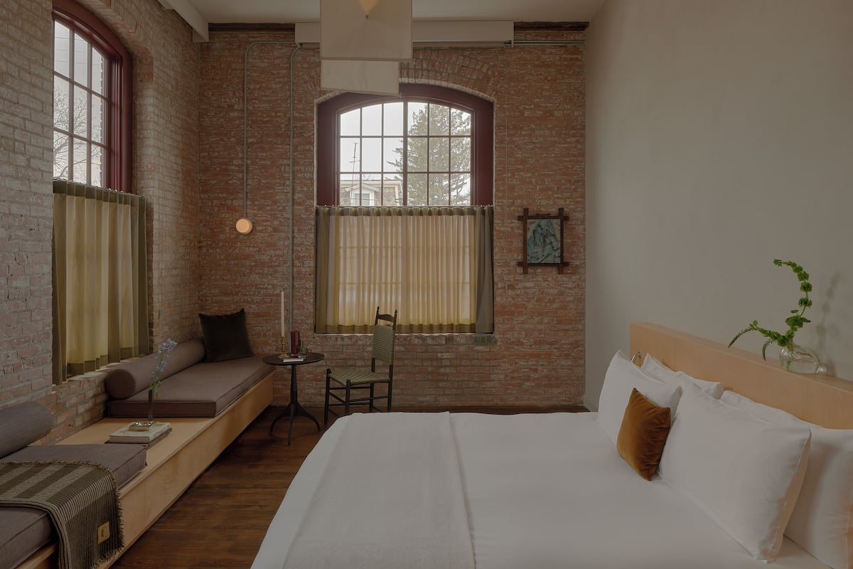 A room with a brick wall, a large arched window, a bed with white linens, a small table with a chair, and a sofa with cushions.