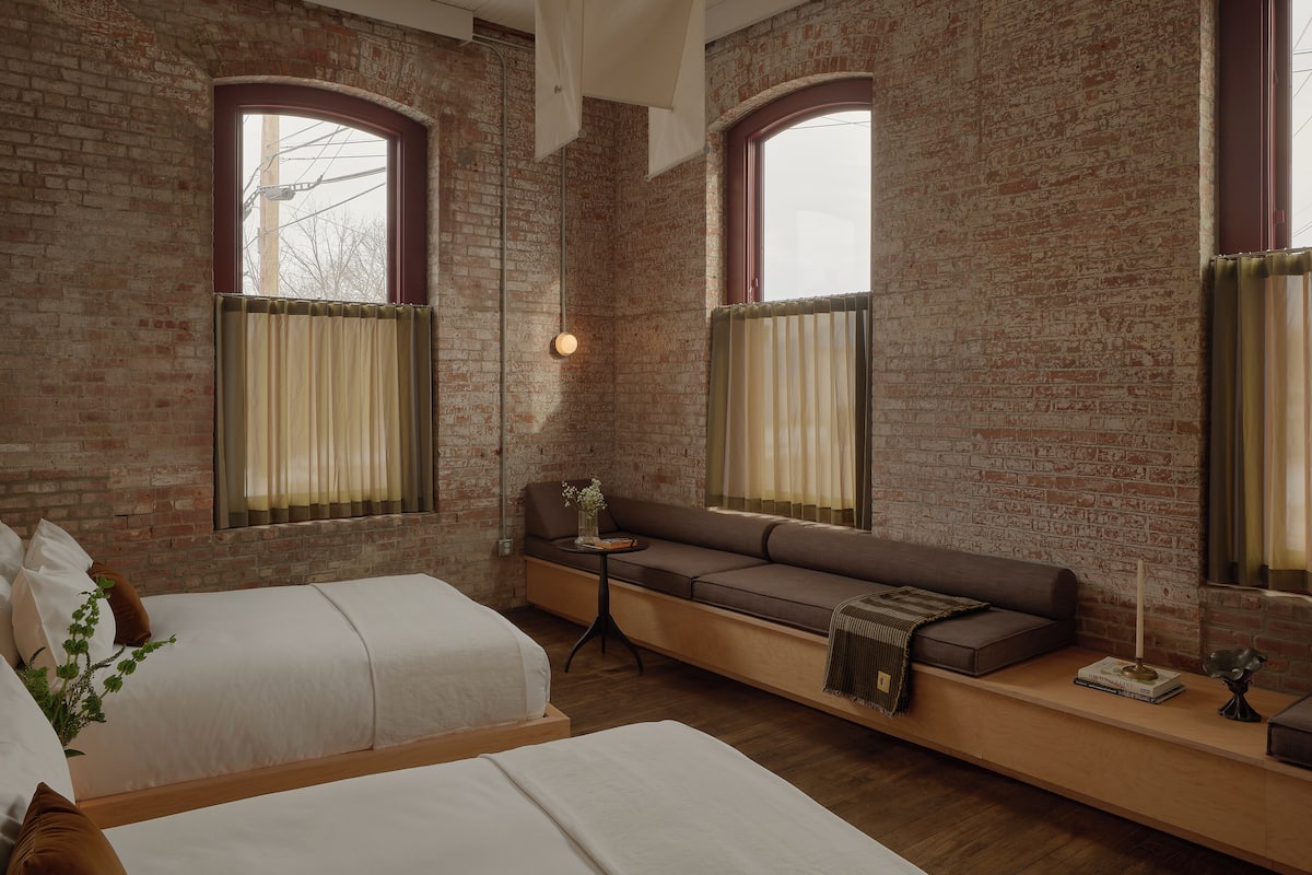 A room with a brick wall, two beds, a bench, and a small table with a lamp.
