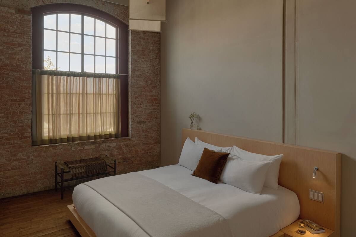 A bedroom with a wooden bed, a bench, a nightstand, a window with curtains, and a brick wall.