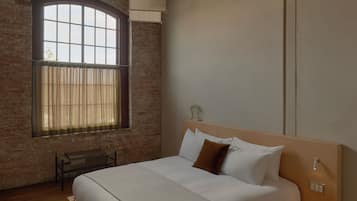 Un dormitorio con una cama de madera, un banco, una mesita de noche, una ventana con cortinas y una pared de ladrillo.