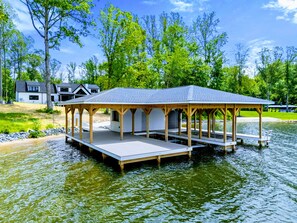 A wooden gazebo with a flat roof and open sides, situated on a dock over water, with a house in the background.