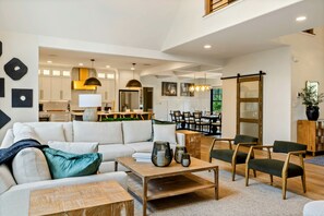A spacious living area with a sofa, coffee table, and chairs. A kitchen with white cabinets and a dining area with chairs and a table are visible in the background.