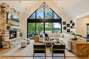 A spacious living room with a stone fireplace, a large window with a view of trees, a modern chandelier, and a sectional sofa. - Lakefront Lake Anna, Beach, Dock, Deck Vie (Bumpass)