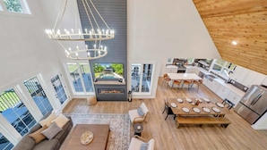A modern interior with a dining area, kitchen, and living space, featuring a fireplace, large windows, and a high ceiling.