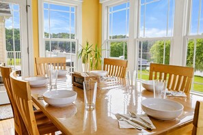 Una mesa de comedor dispuesta con cuencos, vasos y cubiertos, rodeada de sillas de madera, con vistas a la vegetación a través de amplios ventanales.