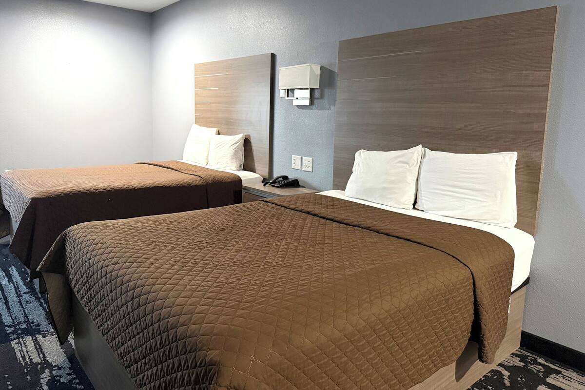 A hotel room with two beds, each with a brown quilted bedspread and white pillows. There is a nightstand with a telephone and a wall-mounted light fixture.