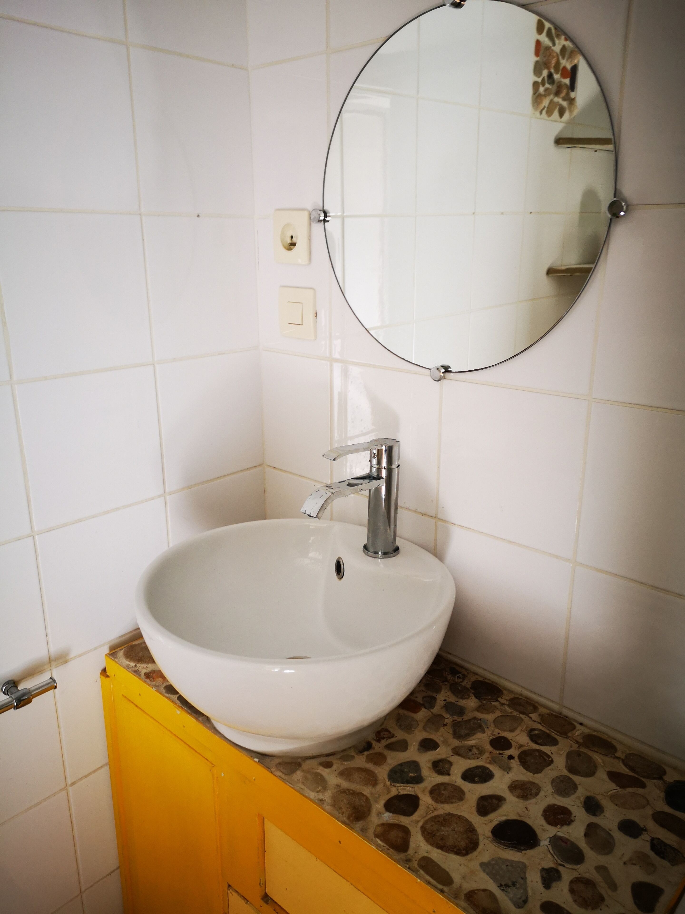 Une salle de bain avec un lavabo blanc, un miroir rond et un meuble jaune.