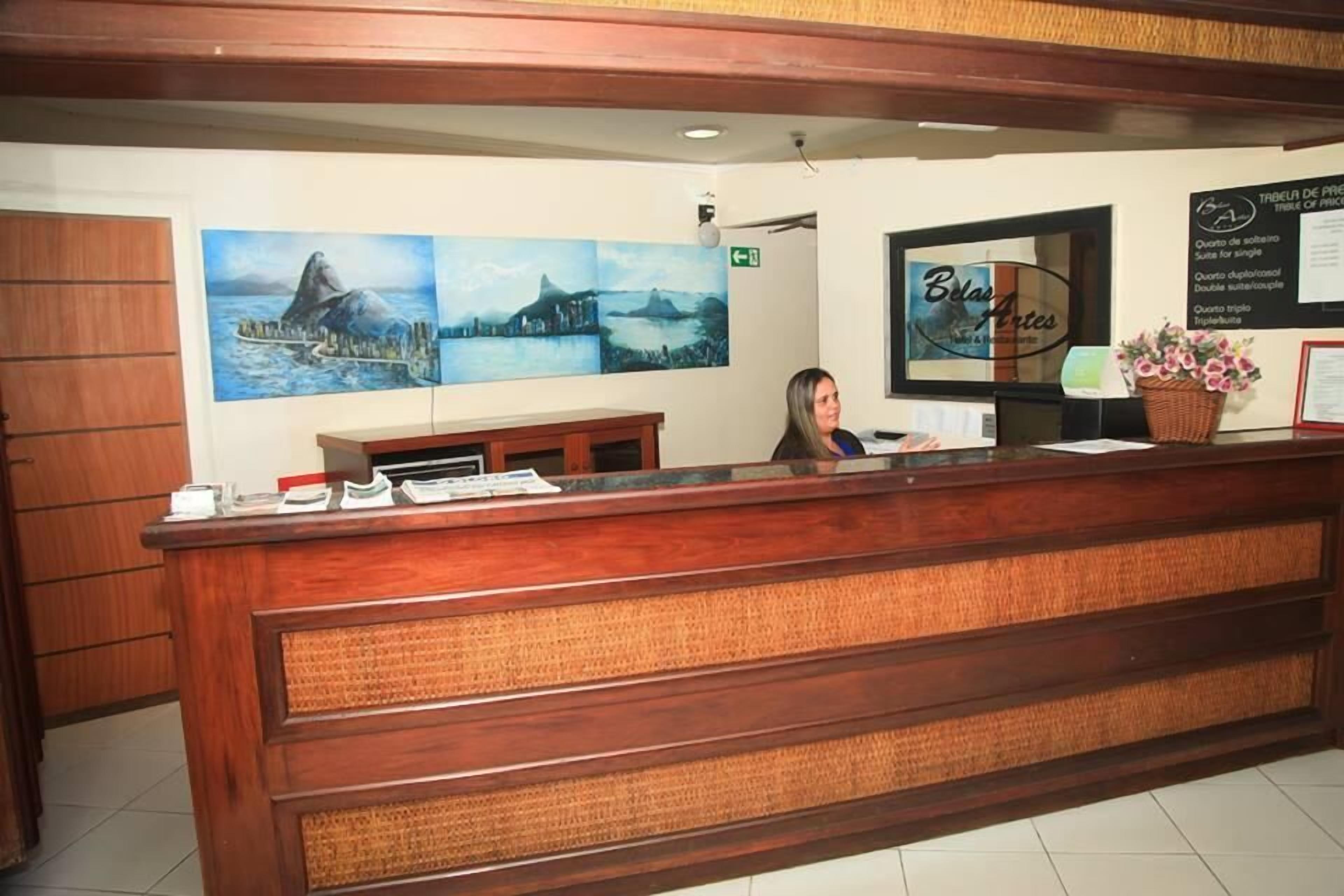 Un comptoir d’accueil avec un revêtement en bois, une femme assise derrière, et trois grandes peintures de paysages accrochées au mur.