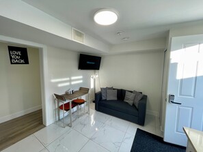 A modern living room with a flat-screen TV, a sofa, a dining table with chairs, and a door with a keypad lock.