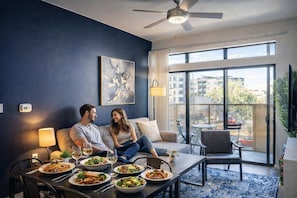 A modern living room with a sofa, dining table, and chairs. There are multiple dishes on the table, and a large window with a view of buildings.