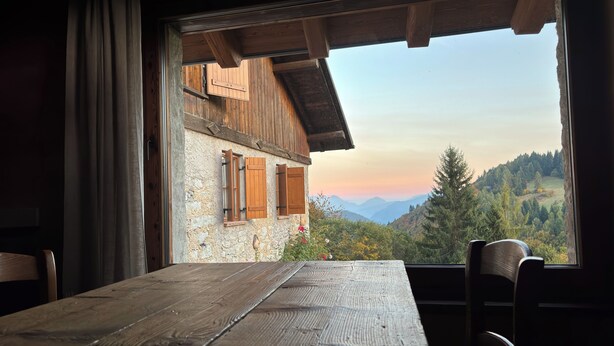 Une table en bois rustique, entourée de chaises, avec vue sur un paysage montagneux par la fenêtre, et un mur de pierres agrémenté de volets en bois.
