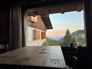 A rustic wooden table with chairs, a view of a mountain landscape through a window, and a stone wall with wooden shutters.