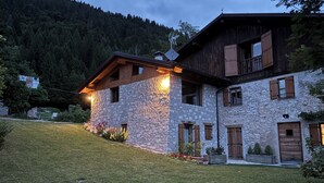 Un bâtiment en pierre, doté de volets en bois et d’un toit en pente, entouré d’une pelouse et d’arbres.