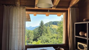 Une table à manger en bois avec un saladier, un buffet en bois contenant de la vaisselle, et une fenêtre avec des rideaux donnant sur un paysage montagneux boisé.