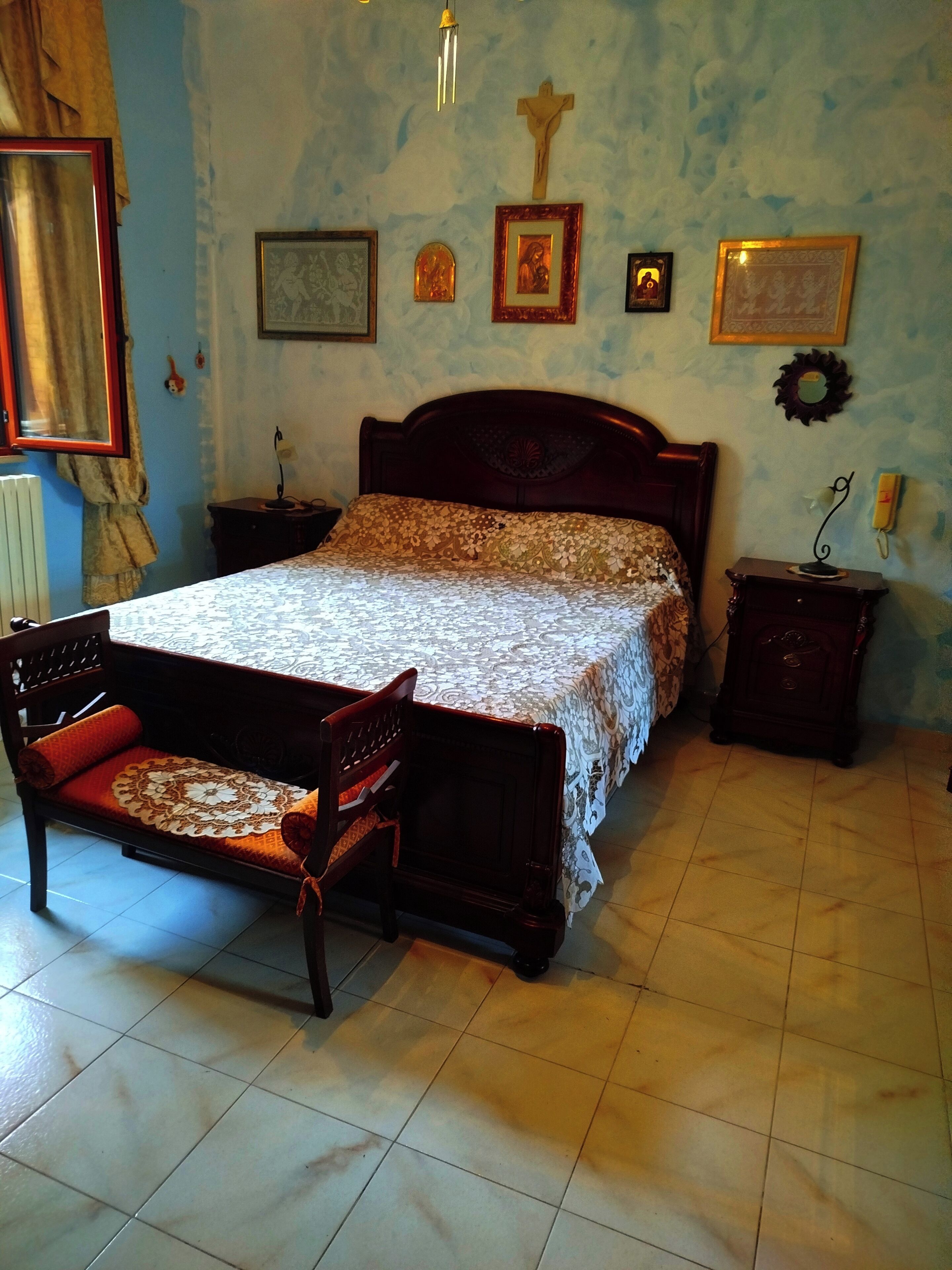 Camera da letto con un letto, una sedia di legno, un comodino, una croce appesa al muro e quadri incorniciati.
