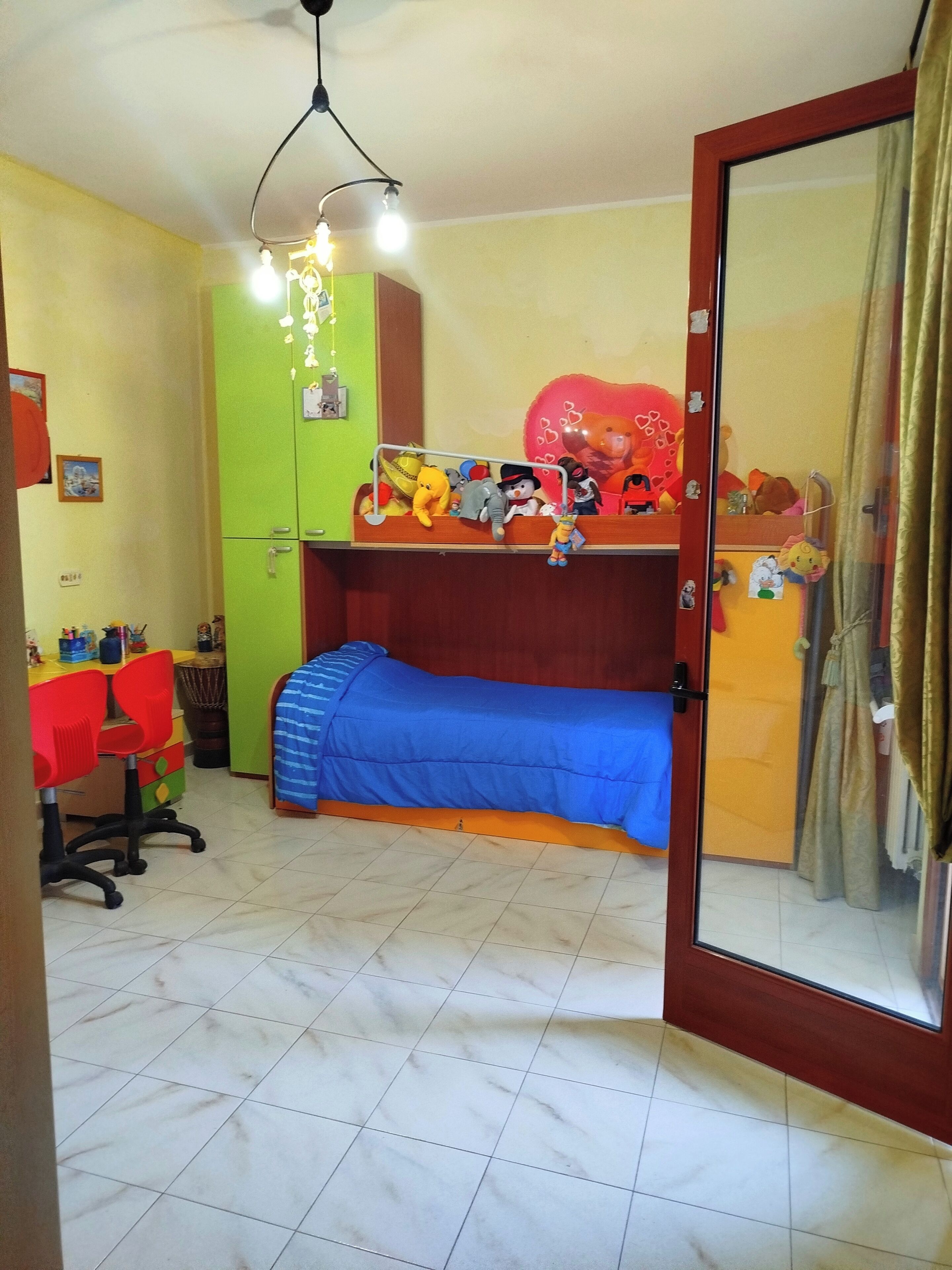 Une chambre avec un lit superposé, un bureau avec une chaise, une armoire verte et une fenêtre avec des rideaux.