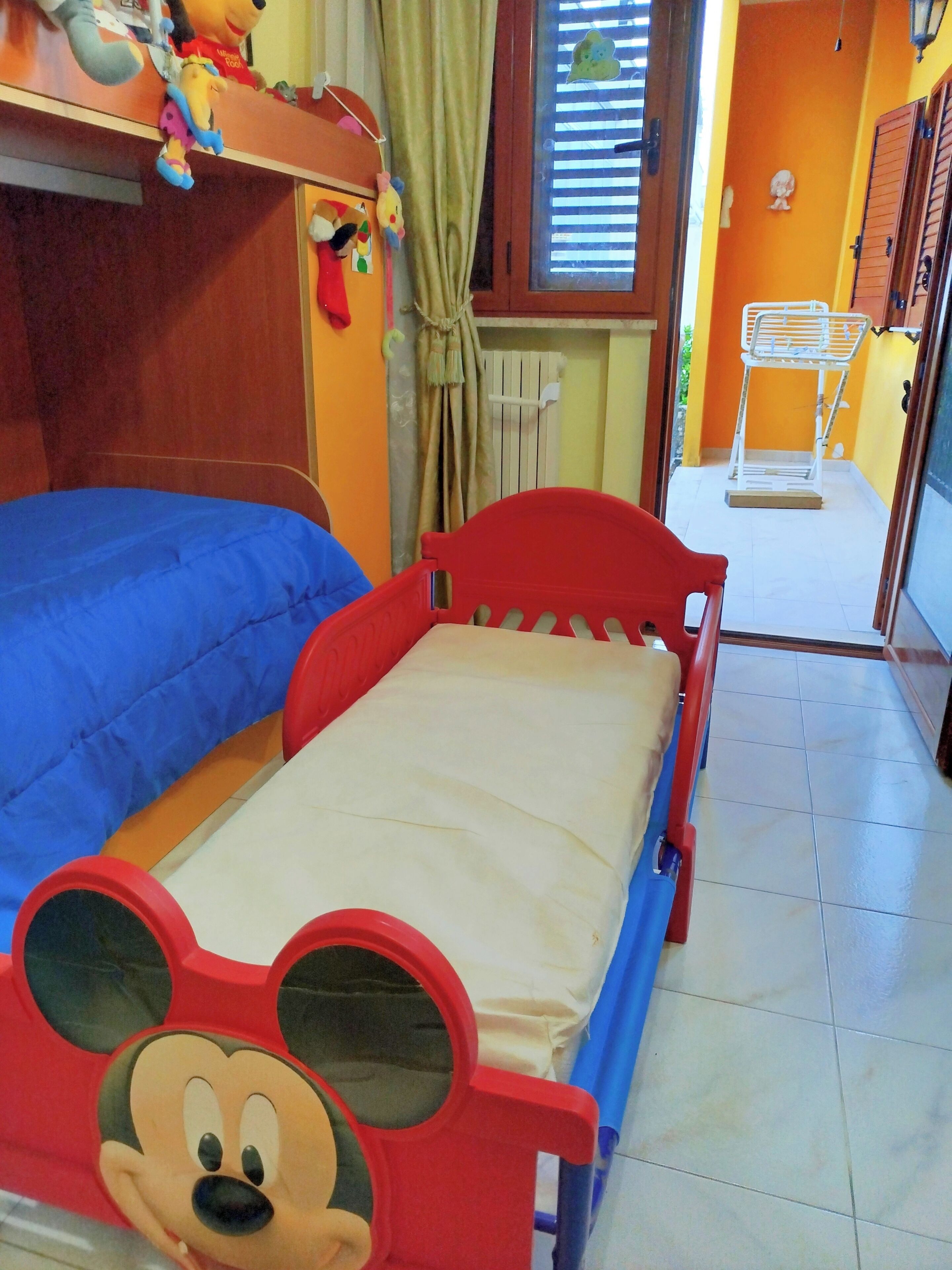 Una stanza con due letti, uno con decorazioni di Topolino e l'altro con copriletto blu. Si vede una finestra con persiane, un termosifone e uno stendibiancheria vicino alla finestra.