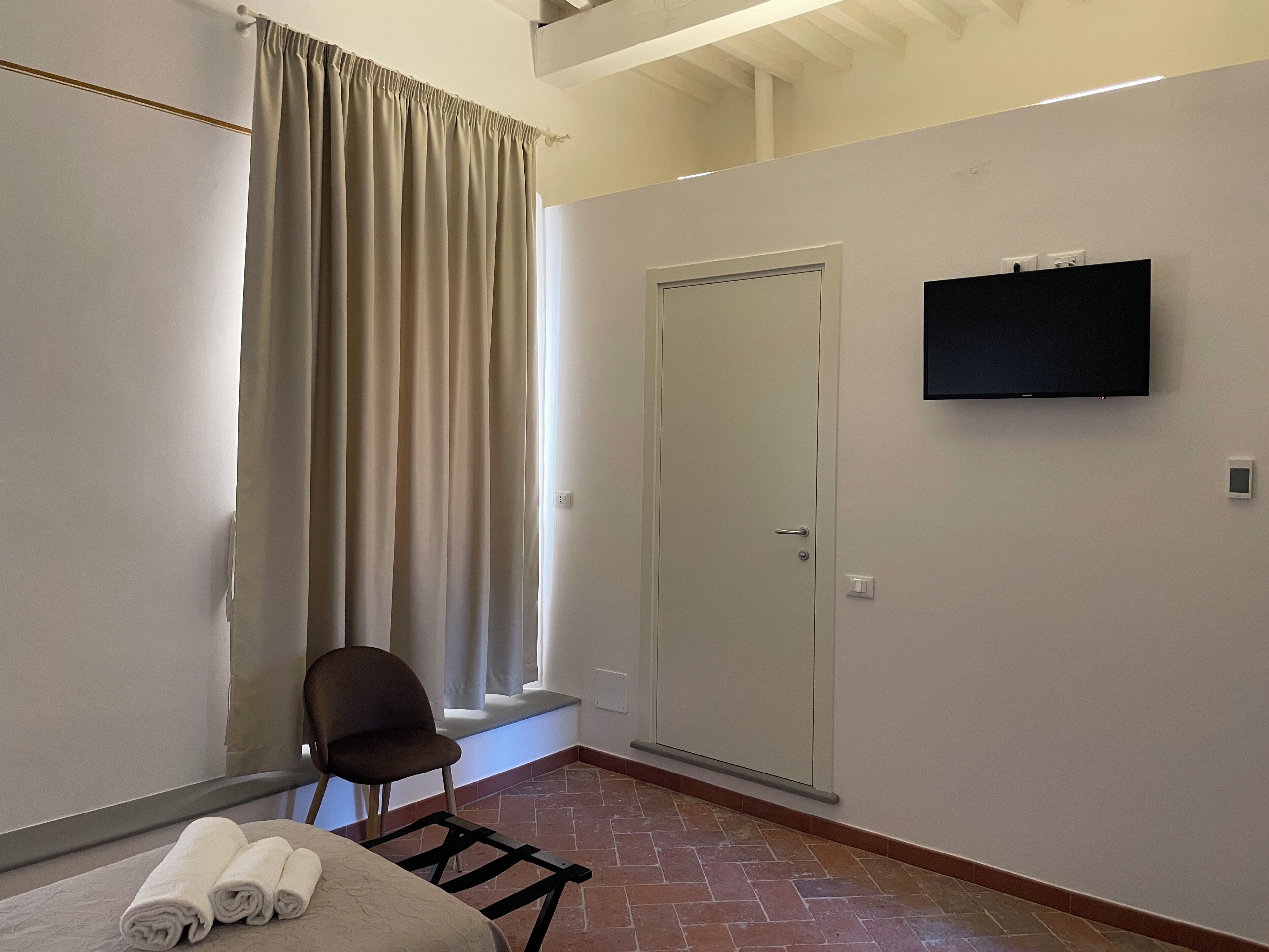 Una camera d'albergo con un letto, una sedia, una televisione, una porta e una finestra con tende.