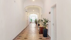 A long, narrow hallway with a tiled floor, white walls, and several doors on one side. There are potted plants along the hallway, and a fire extinguisher is visible in the distance. - IL GENIO Residenza d'Epoca (Pisa)