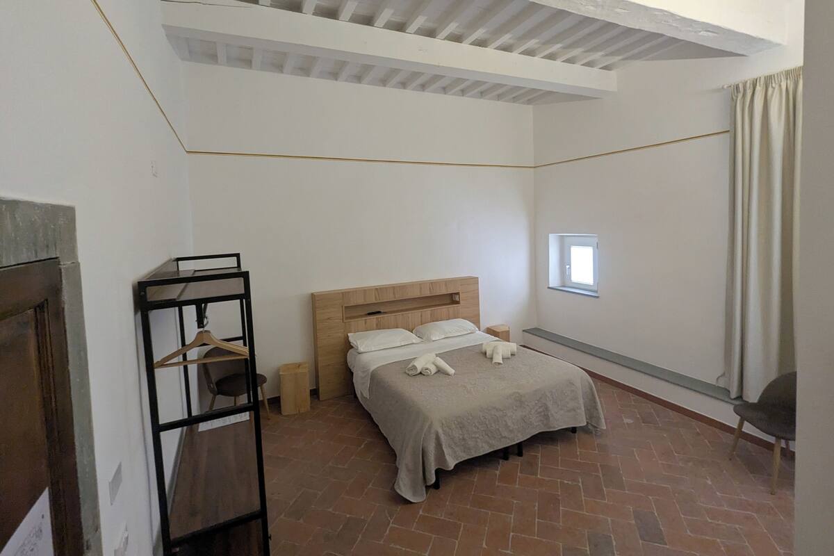 A bedroom with a bed, a small window, a wooden headboard, a hanging rack, and a brick floor.