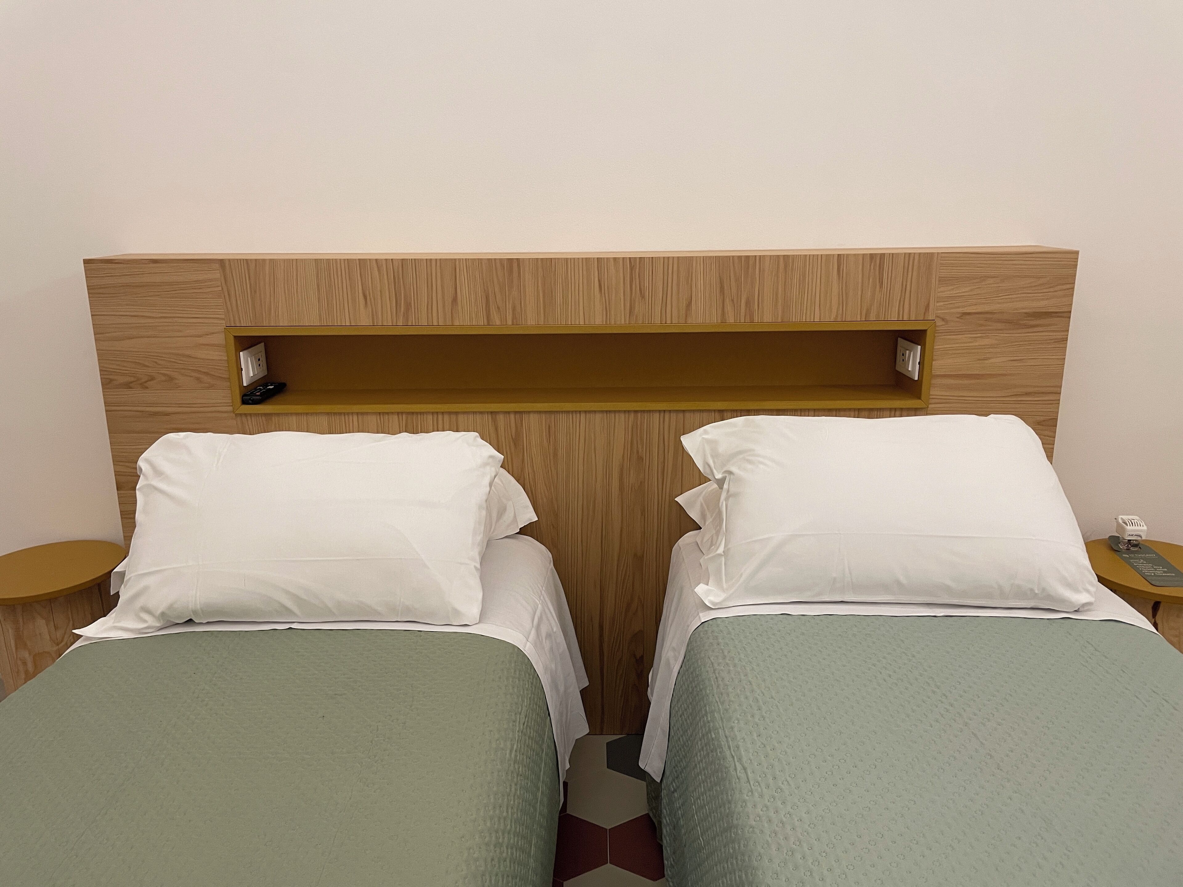 Een hotelkamer met twee eenpersoonsbedden, een houten hoofdbord, een klein plankje en een nachtkastje met telefoon en oplader.