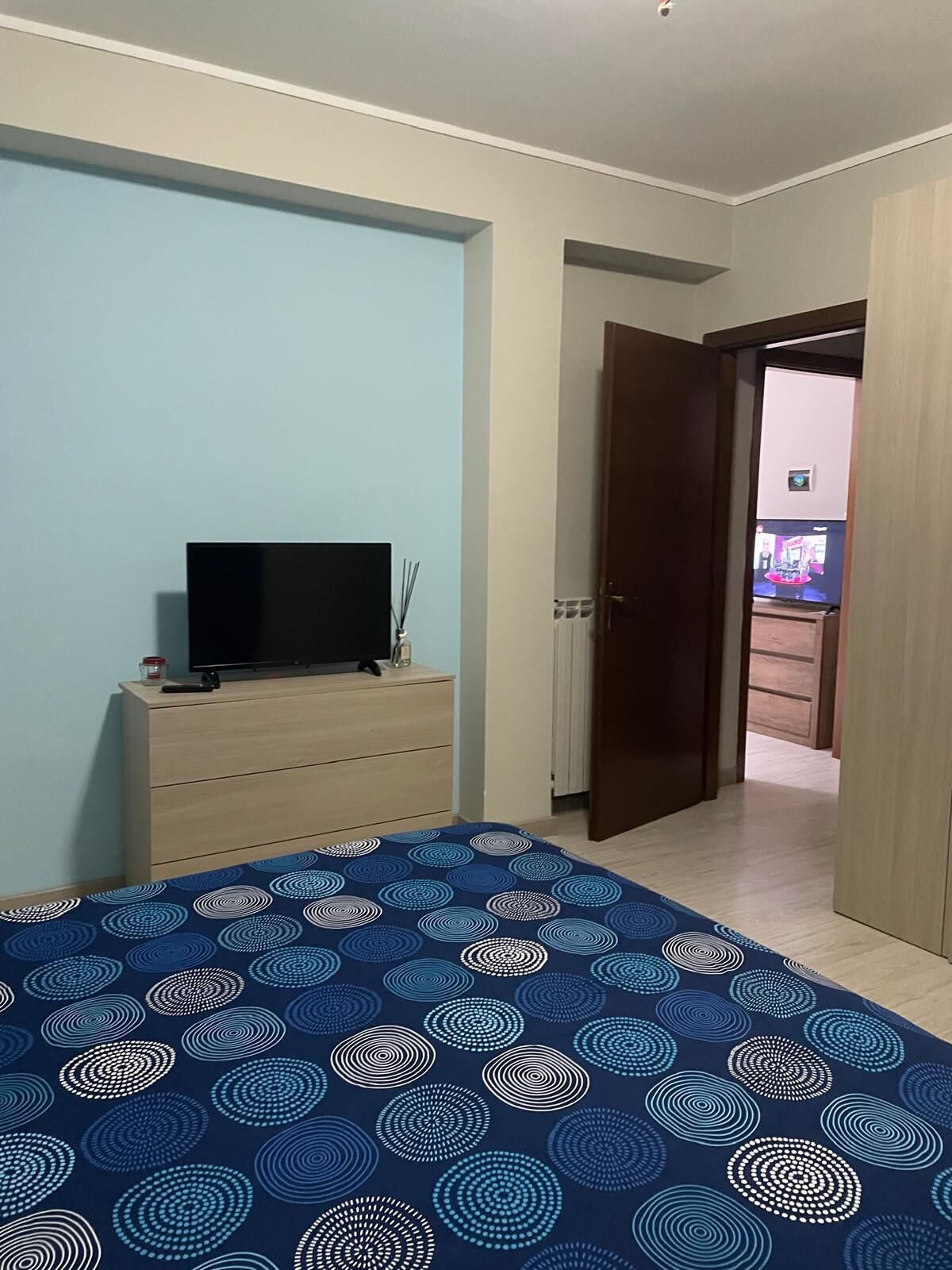 Un dormitorio con cama, televisor sobre una cómoda, puerta que da a otra habitación y edredón estampado.