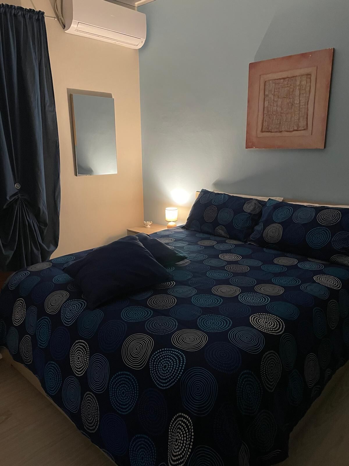 A bedroom with a bed, a nightstand, a lamp, a wall-mounted air conditioner, and a framed picture.