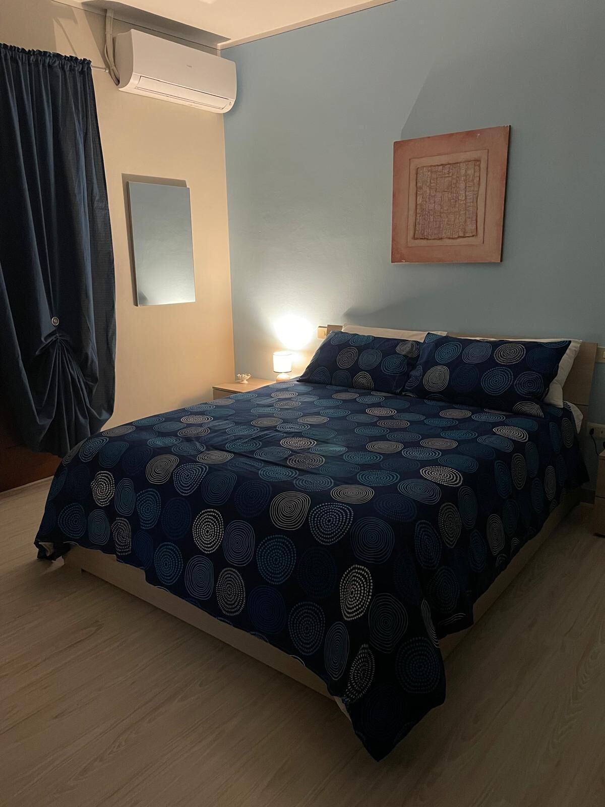 A bedroom with a bed, a nightstand, a wall-mounted air conditioner, a window with curtains, and a framed picture on the wall.