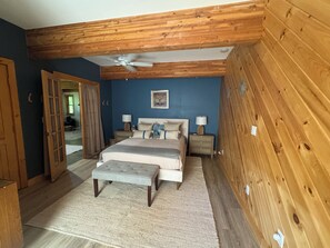 Ein Schlafzimmer mit Holzboden, einem Bett mit grauem Polsterkopfteil, zwei Nachttischen mit Lampen und einem großen Fenster.