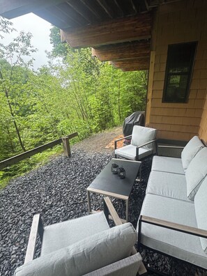Et overdækket udendørs opholdsområde med sofa, stole og et bord, omgivet af grus og grøn vegetation.