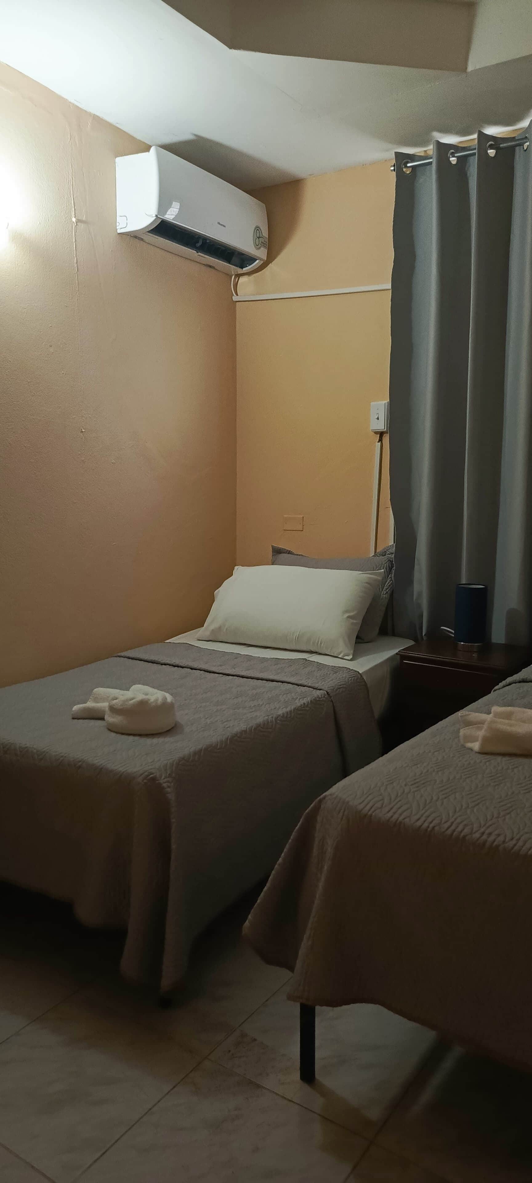 Una habitación de hotel con dos camas individuales, un ventilador de techo, un equipo de aire acondicionado, una mesita de noche con lámpara y una ventana con cortinas.