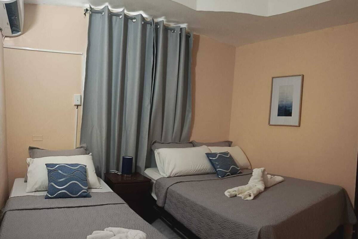 A hotel room with two single beds, a bedside table with a lamp, a framed picture on the wall, and a window with grey curtains.