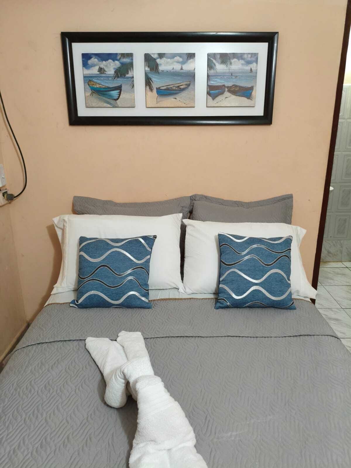 A bed with a grey bedspread and two pillows with a blue and white wavy pattern. Above the bed, there is a framed picture of boats on a beach.