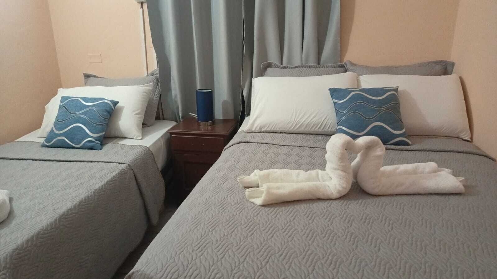 Camera d'albergo con due letti, ciascuno con copripiumino grigio e cuscini coordinati. Tra i letti si trova un comodino con una lampada.