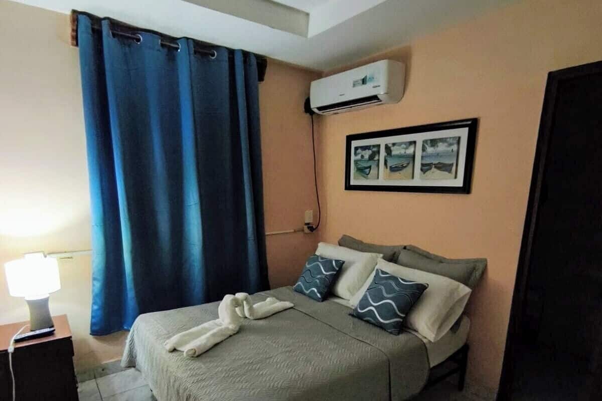 A bedroom with a bed, a nightstand, a lamp, a wall-mounted air conditioner, and a window with blue curtains.
