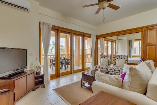 A spacious living room with a flat-screen TV, wooden furniture, a ceiling fan, and large sliding glass doors leading to an outdoor patio.