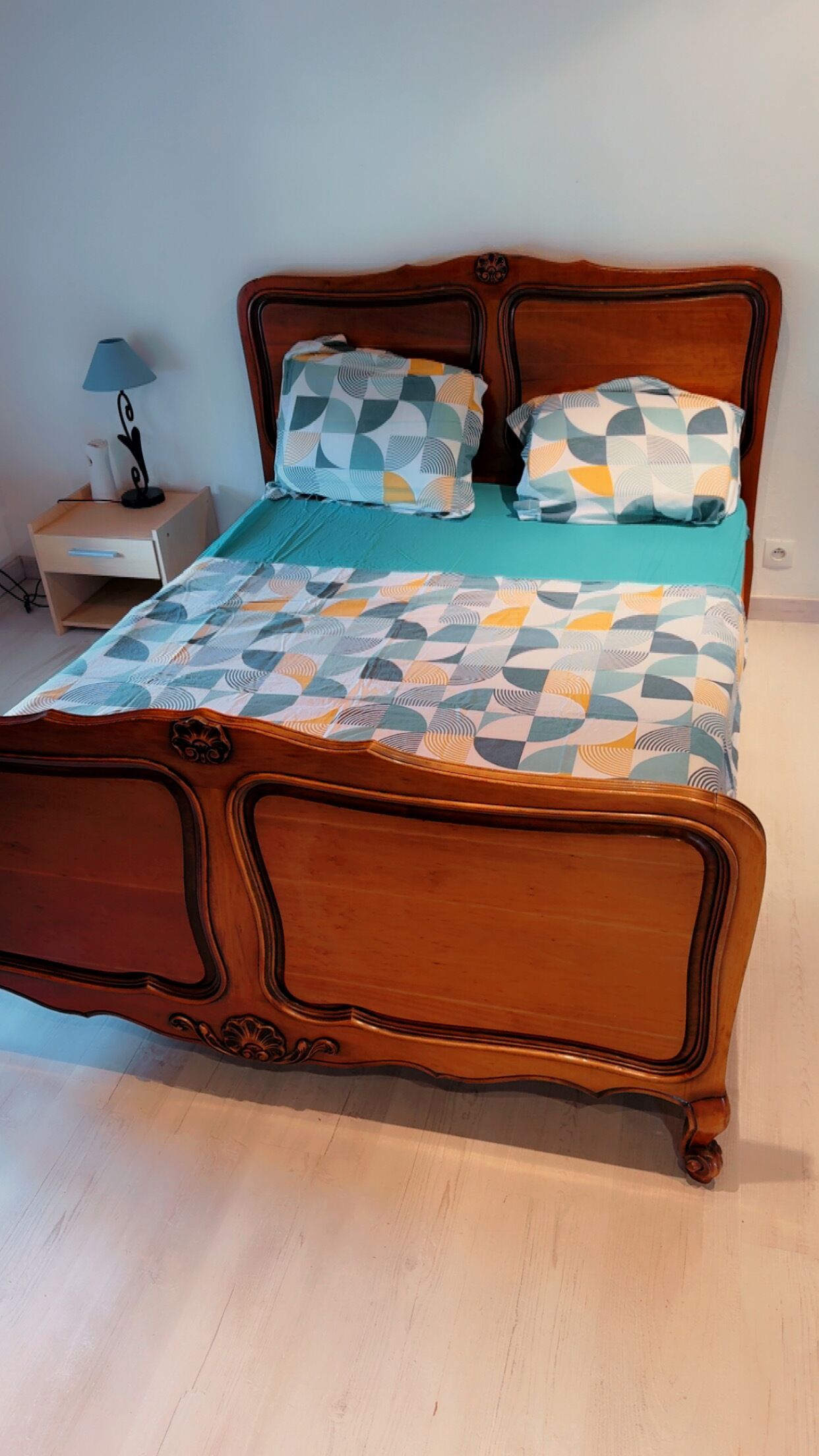 A wooden bed with a geometric patterned bedspread and pillows, a bedside table with a lamp, and a nightstand.