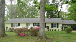 A single-story house with a gabled roof, a chimney, and a well-maintained lawn with flowering shrubs.