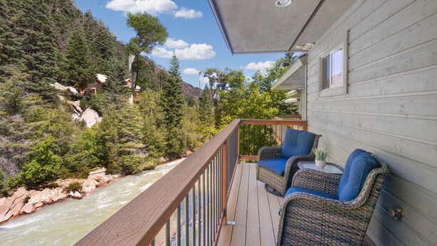 Terraza de madera con mobiliario de mimbre con vistas a un río y montañas boscosas.