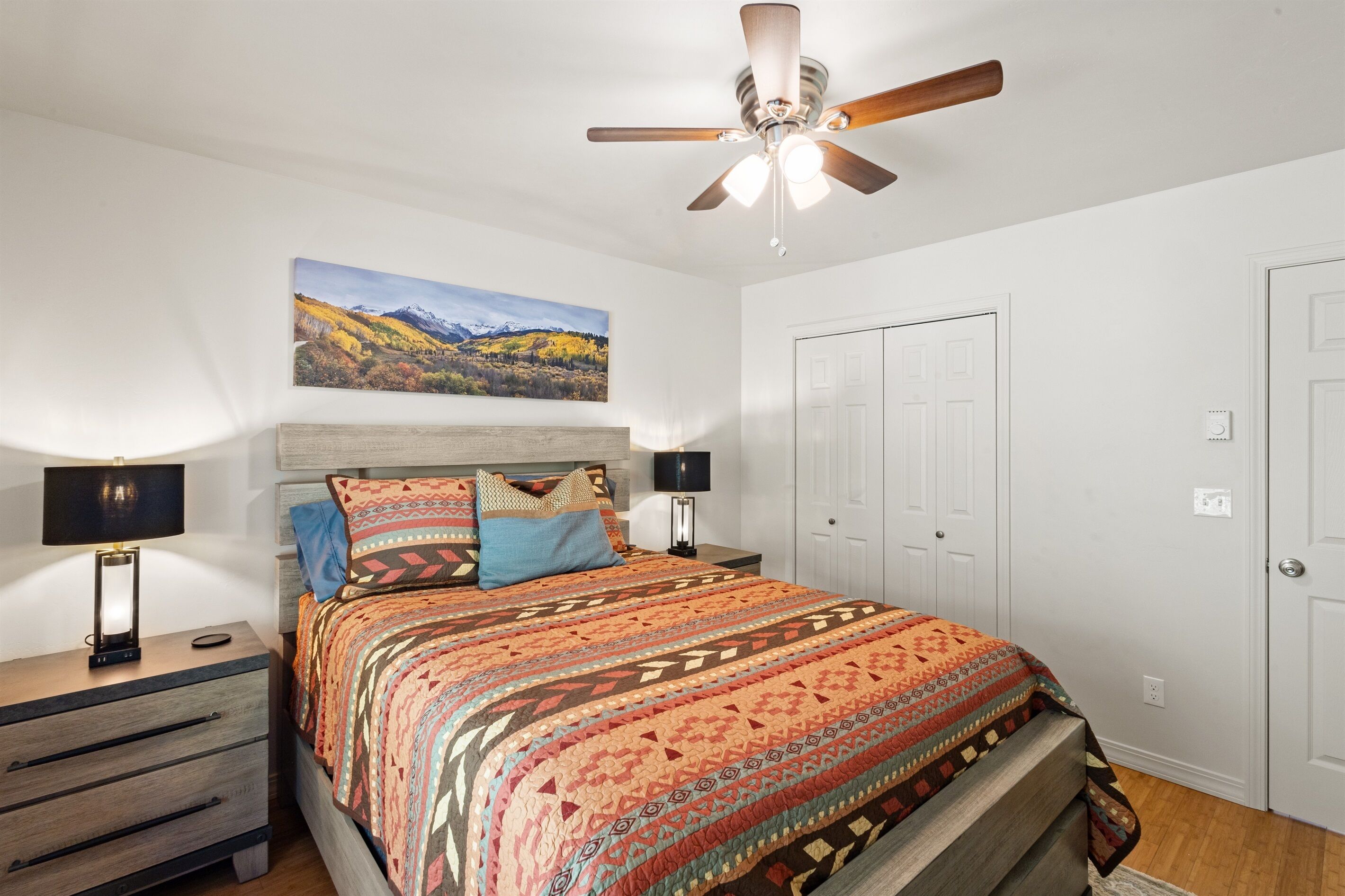 A bedroom with a bed, a ceiling fan, a nightstand with a lamp, a wall-mounted painting, and a door.