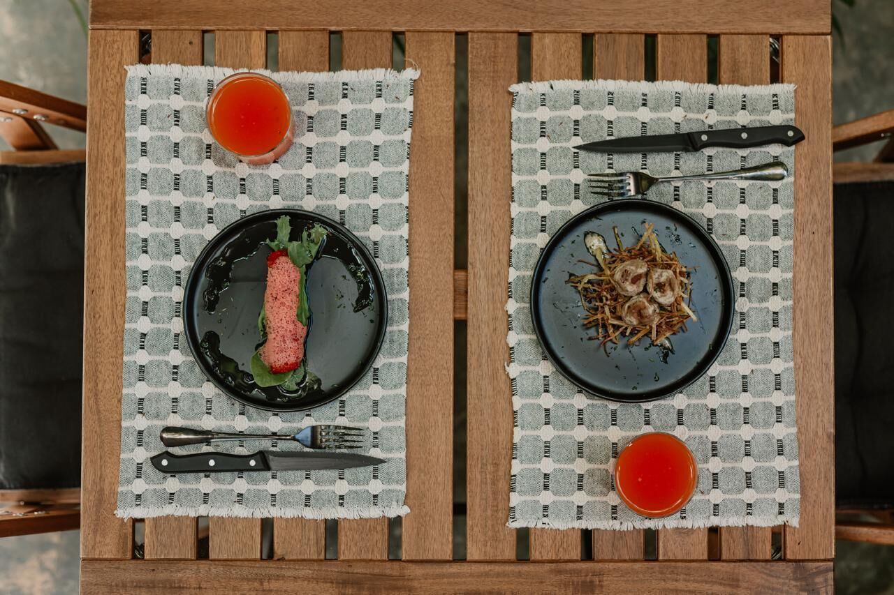 A wooden table with two plates of food and two glasses of red liquid, possibly juice or sauce, on a checkered tablecloth.