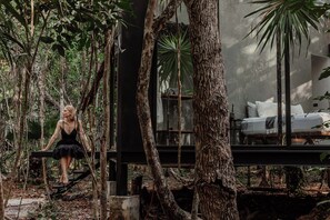 A woman sitting on a wooden bench in a forested area, overlooking a bed with white linens and pillows in a glass-walled room. - SOTA Human Space Retreat Centre & Wellness Adults Only (Puerto Morelos)