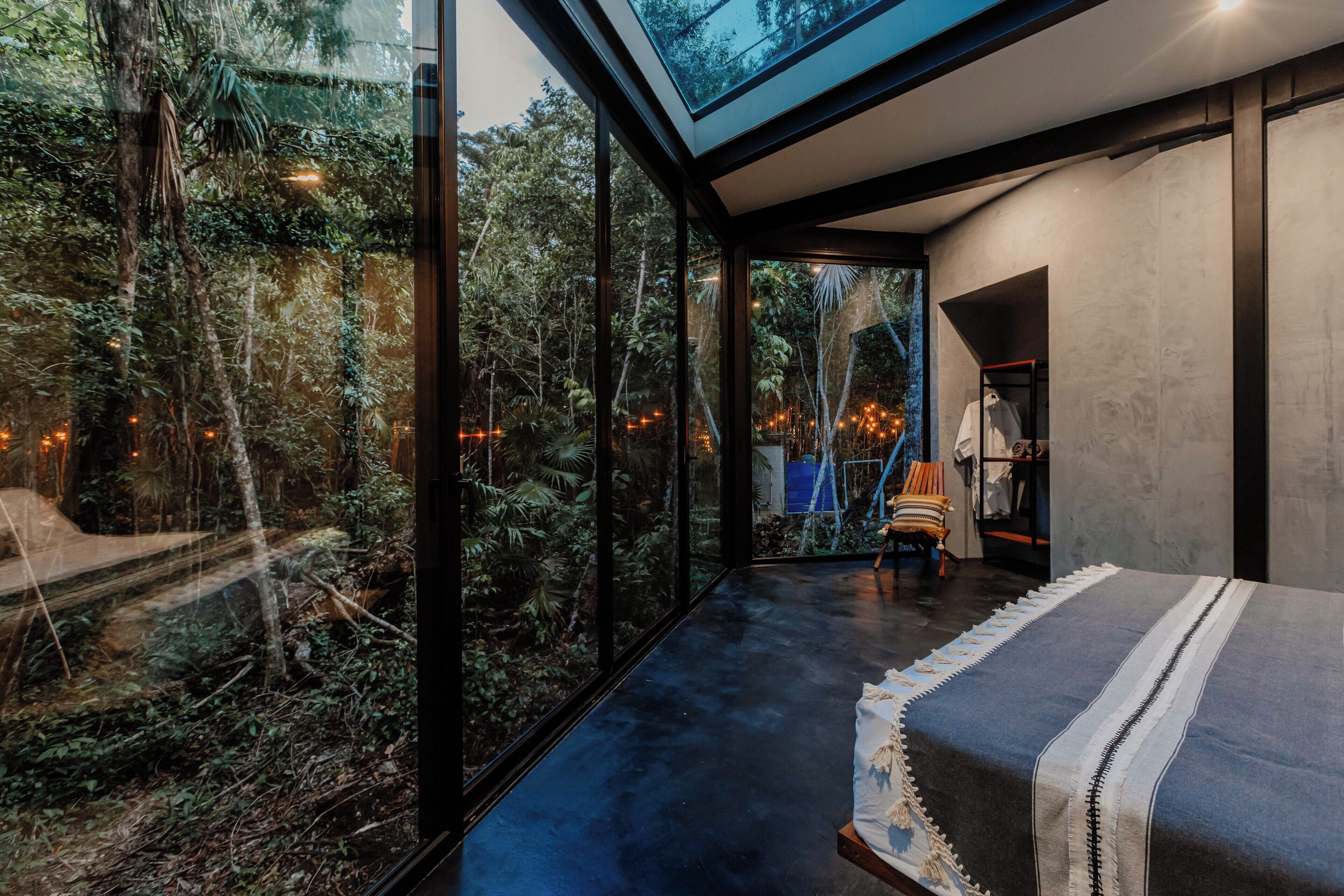 Ein modernes Schlafzimmer mit großen Glasfenstern, einem Bett mit gestreifter Decke und Blick auf eine bewaldete Landschaft.