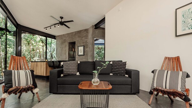 A modern living room with a dark sofa, two wooden chairs, a wooden coffee table, and a ceiling fan. - SOTA Human Space Retreat Centre & Wellness Adults Only (Puerto Morelos)