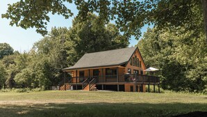 Une cabane en rondins dotée d’un balcon, entourée d’arbres et d’une zone herbeuse.