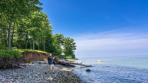 Un rivage rocheux où se trouve une personne, debout près du bord de l’eau, entourée d’arbres et sous un ciel dégagé.