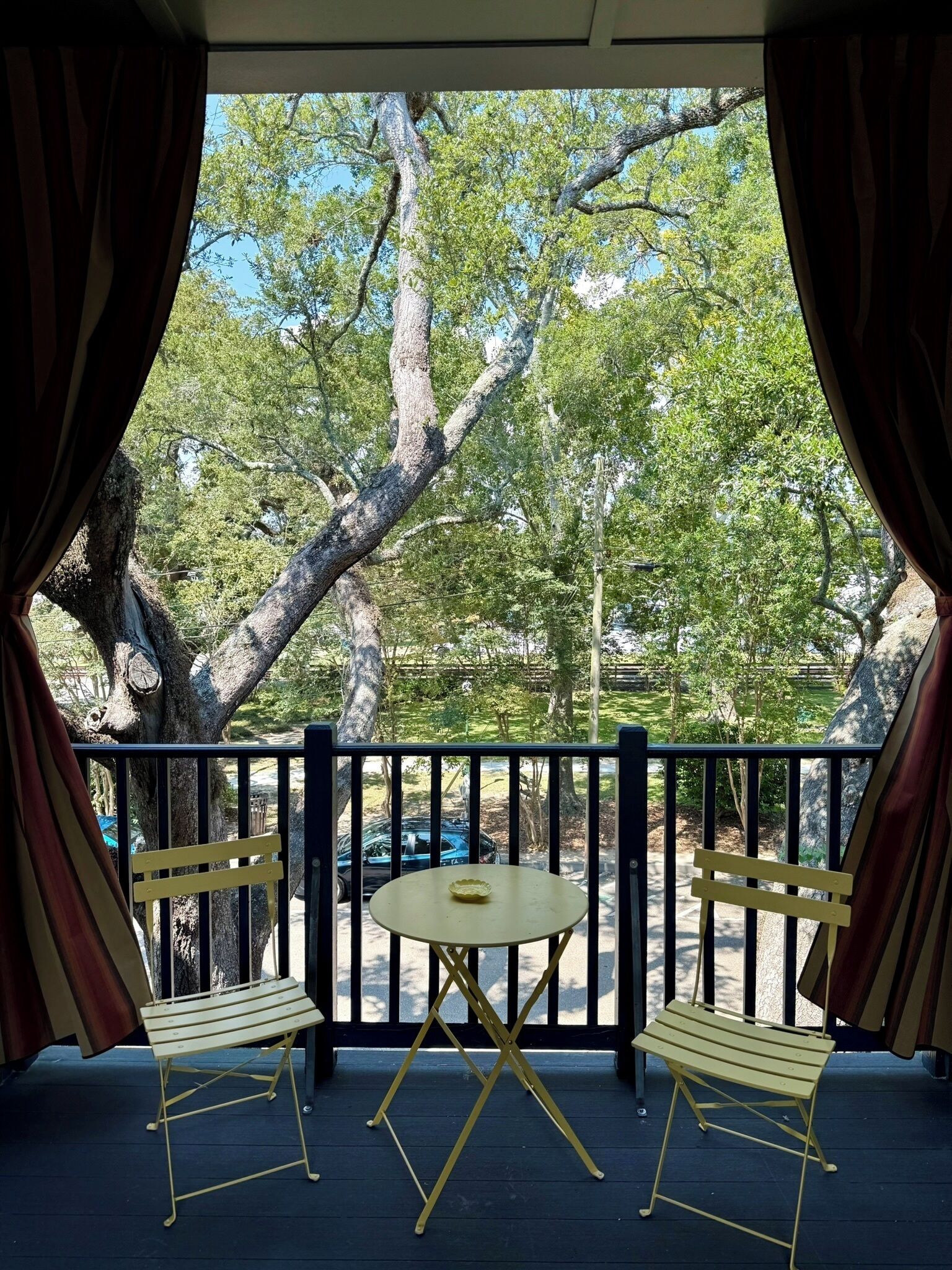 A covered outdoor seating area with a table and two chairs, a railing, and a view of trees and a car in the background.