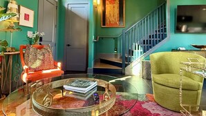 Una sala de estar con paredes color turquesa, una mesa de centro de vidrio, un sillón rojo, un sillón verde, una escalera y un televisor montado en la pared.