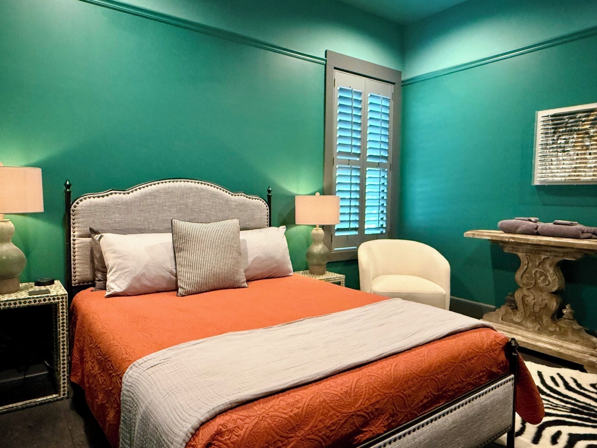 Un dormitorio con cama, mesitas de noche, una silla, una mesa y una ventana con contraventanas.