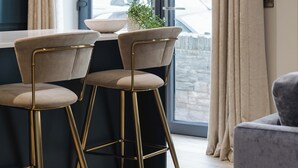 A modern kitchen with a dining area, featuring a large window with curtains, a potted plant, and two bar stools. - Experience a sense of deep calm with seaside walks & blissful sea air (Portrush)